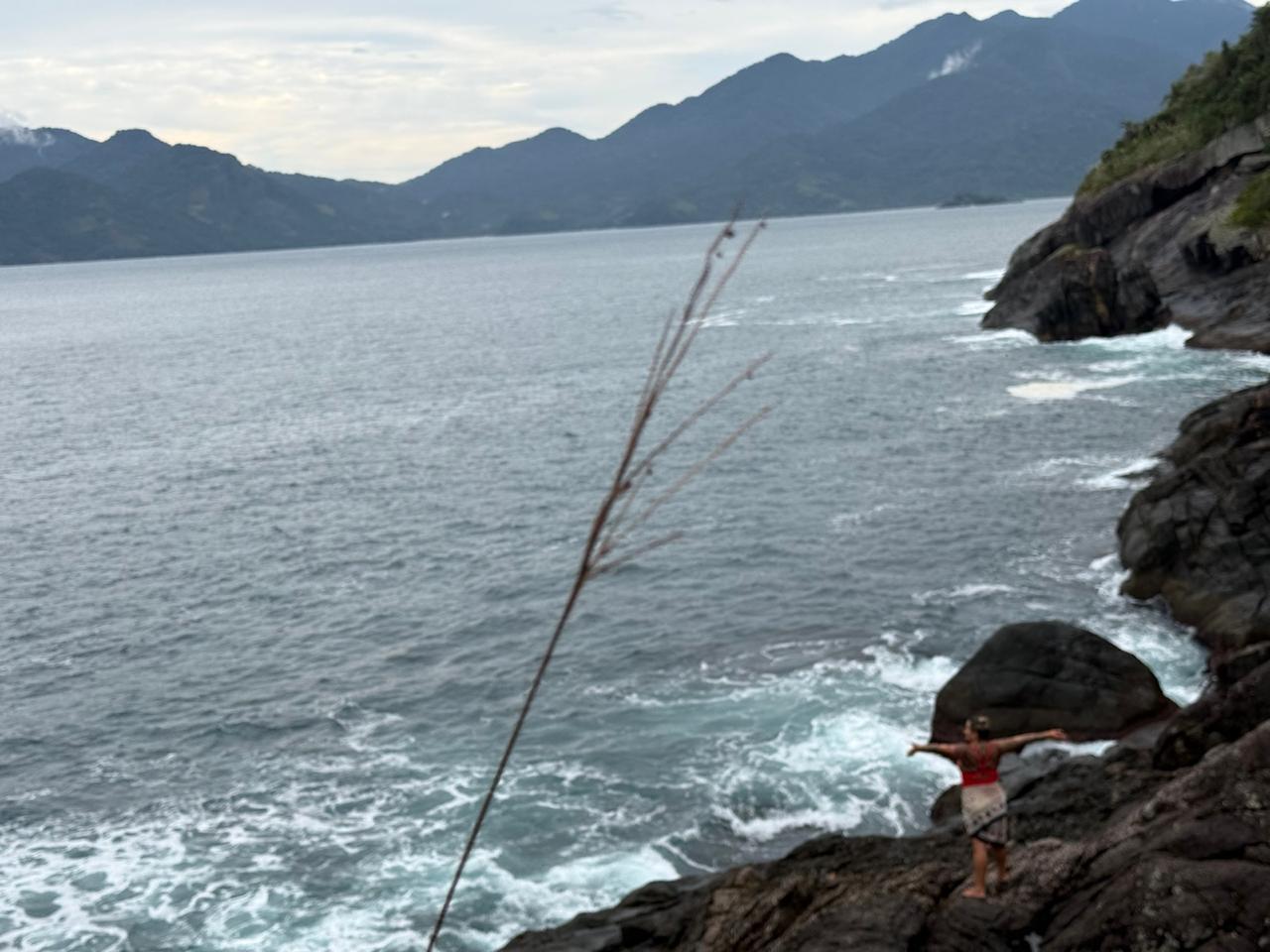 Passageira em pé sobre pedras à beira do costão com braços abertos, mar batendo forte e montanhas ao fundo
