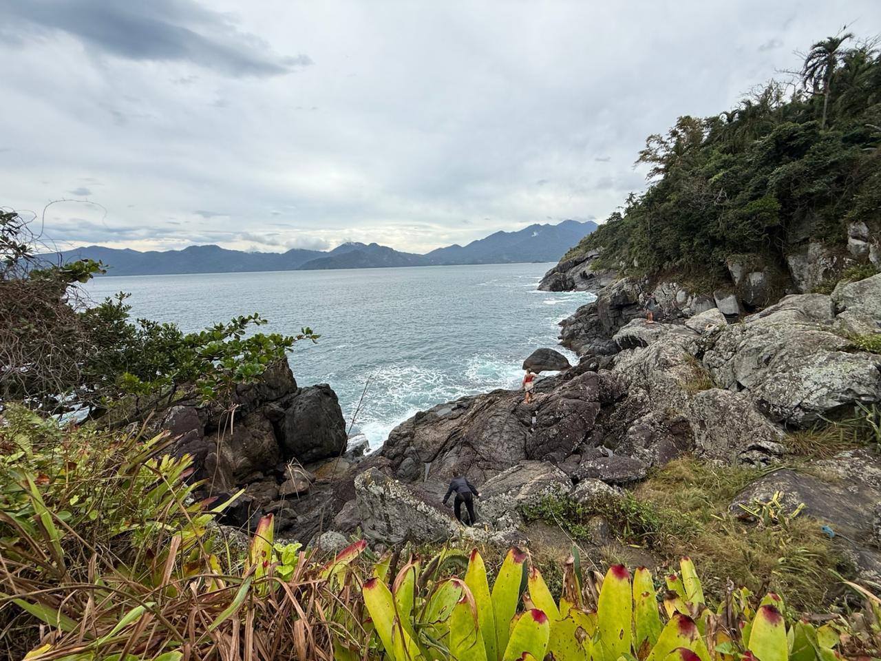 Grupo em trilha pelo costão rochoso com bromélias em primeiro plano, vista para o mar de Ilhabela
