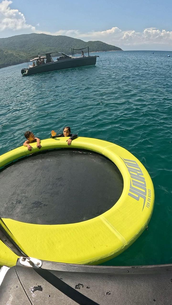 Criancas brincando na boia inflavel Yourou com a lancha ancorada ao fundo em Ilhabela