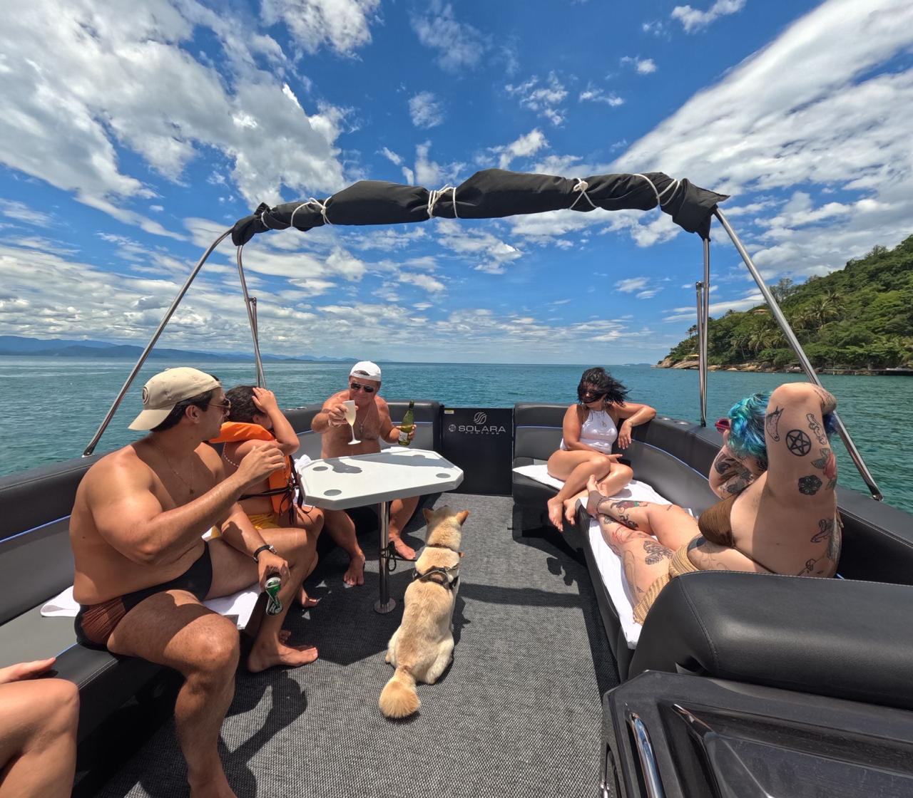 Passageiros no deck da lancha em dia ensolarado, praia e mata ao fundo