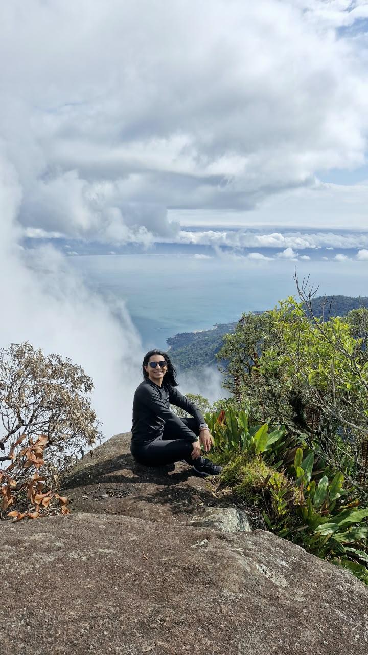Aventureira sentada em pedra no topo do Pico do Baepi com nuvens ao lado - sensacao de topo do mundo